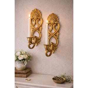 May include: Two ornate gold wall sconces with lit white candles. The sconces have a shell-like design at the top and an intricate, swirling pattern. The sconces are mounted on a textured wall. The overall theme is home decor.