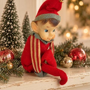 May include: A vintage-style elf figurine wearing a red felt hat with a grey brim, a matching red and grey collar, and a red suit with cream-colored vertical stripes on the sleeves. The elf has a painted face with blue eyes and pointed ears.