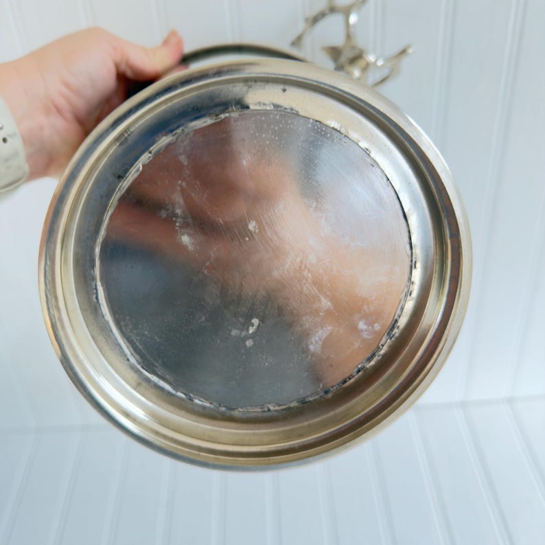 May include: A silver-colored metal serving tray with a circular design. The tray has a reflective surface and a decorative handle. The bottom of the tray shows signs of wear and use.