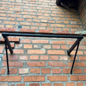 Puede incluir: Una barra de dominadas de metal negro montada en una pared de ladrillo rojo. La barra es horizontal, con soportes angulados. La albañilería presenta tonos de rojo y marrón.