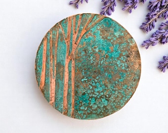 May include: A round, copper-colored decorative piece with a tree design. The tree trunk and branches are a lighter copper color, set against a mottled turquoise background. Sprigs of purple flowers are in the background.