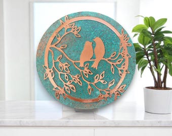 May include: A round copper wall art piece with a turquoise background. The art features two birds perched on a branch with leaves. The birds and branch are cut out of the copper and are silhouetted against the turquoise background.