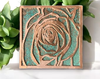 May include: A square copper tile with a raised design of a rose in a teal blue color.
