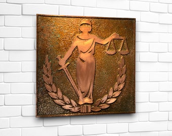 May include: A copper-toned wall plaque featuring Lady Justice, blindfolded, holding scales and a sword. The figure is surrounded by a laurel wreath, set against a textured, square background. The artwork is mounted on a white brick wall.