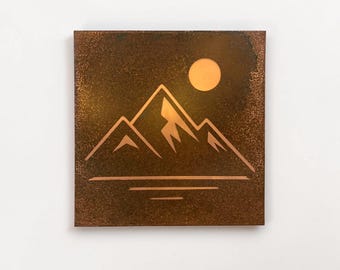 May include: Square copper-colored wall art featuring a mountain range silhouette with a sun above and horizontal lines below. The artwork has a textured surface and is mounted on a white wall.