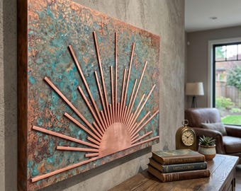 May include: A rectangular wall art piece featuring a copper sunburst design with radiating rays mounted on a textured background of oxidized copper with mottled blue, green, and brown tones. The sunburst is made of polished copper metal.