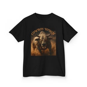 May include: Black t-shirt featuring a graphic of a charging bull with the text "Cowboy Tough" in orange. The bull is depicted in a cloud of dust, conveying a sense of power and action. The design is centered on the shirt.