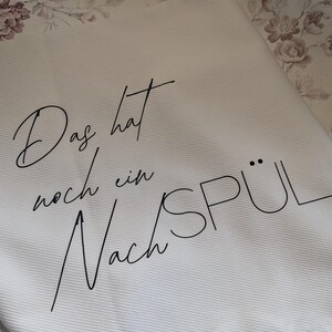 Könnte beinhalten: Weißes Küchentuch mit schwarzer Schrift, die "Das hat noch ein Nachspül" lautet. Das Handtuch ist gefaltet und auf einer Oberfläche mit floralem Muster platziert. Das Handtuch besteht aus einem strukturierten Stoff.