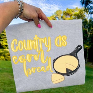 Könnte beinhalten: Ein graues Küchentuch mit dem gelben Schriftzug "Country as cornbread". Eine Cartoon-Illustration von Maisbrot in einer gusseisernen Pfanne und eine Scheibe Maisbrot sind ebenfalls abgebildet.