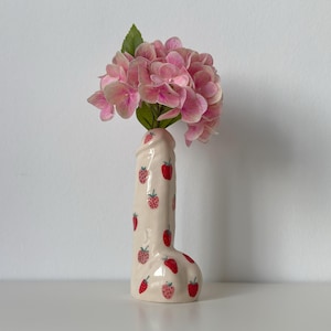 Peut inclure: Un vase en céramique en forme phallique avec un vernis de couleur crème décoré de petits motifs de fraises rouges. Le vase contient un bouquet de fleurs d'hortensia roses avec des feuilles vertes dépassant de l'ouverture supérieure.
