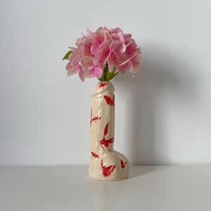Pode incluir: Um vaso de cerâmica cor de creme em forma fálica decorado com pequenas ilustrações de borboletas vermelhas. O vaso contém um cacho de flores de hortênsia rosa vibrante, dispostas na abertura superior da estrutura de cerâmica vertical.