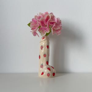 Könnte beinhalten: Eine phallisch geformte Keramikvase mit glänzender cremefarbener Glasur, verziert mit einem wiederkehrenden Muster aus kleinen roten Erdbeeren. Die Vase enthält ein Bündel hellrosa Hortensienblüten mit grünen Blättern an der Basis.