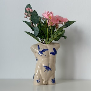 Puede incluir: Un jarrón de cerámica color crema con forma de torso masculino, adornado con diseños de mariposas azules. El jarrón contiene flores rosas y follaje verde. El jarrón es un artículo decorativo.