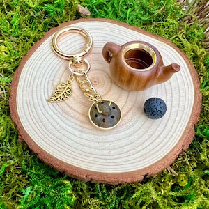 May include: A gold-colored keychain with a leaf charm and a round, stone-like pendant. A small, wooden teapot and a black, porous ball sit on a wooden slice, surrounded by green moss.
