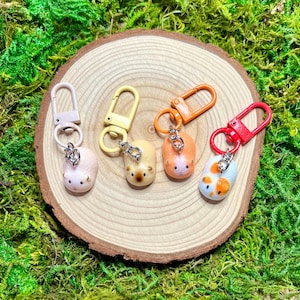 May include: Four small hamster-shaped charms attached to colorful lobster claw clasps. The charms are light pink, cream with a brown nose, light orange, and white with orange spots. Each charm features a silver-tone metal jump ring connector.