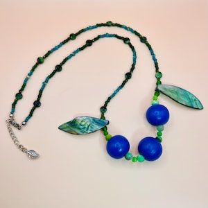 May include: A handcrafted necklace featuring a combination of blue, green, and silver elements. The necklace includes blue wooden beads, green beads, and iridescent leaf-shaped pendants. The clasp has a leaf charm.