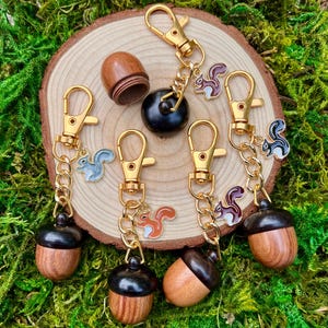 May include: A collection of keychains featuring wooden acorn pendants with gold-colored hardware. Each keychain has a small, colorful squirrel charm. The keychains are arranged on a wooden slice, surrounded by green moss.