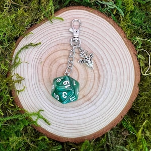 May include: A green, 20-sided dice keychain with a silver dragon charm. The dice has white numbers on each side. The keychain is hanging from a silver chain.