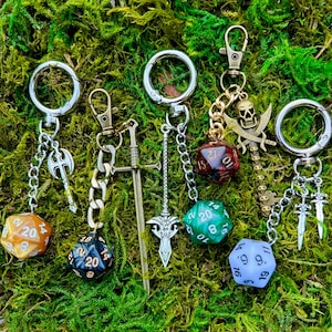 May include: Five metal keychains featuring polyhedral twenty-sided dice in orange, black, green, red, and light purple, each attached to silver or bronze-toned charms including axes, swords, and a skull-topped key with crossed blades.