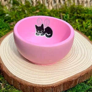 May include: A small, pink, round bowl with a black cat illustration with fairy wings. The bowl is sitting on a wooden slice, with a mossy green background.