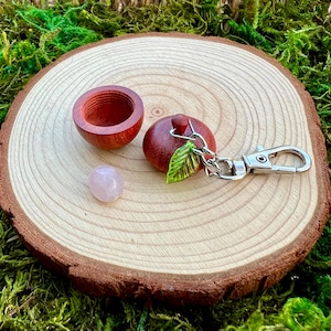 May include: A small, hollow, reddish-brown wooden container shaped like an acorn, featuring a matching lid with a silver-toned metal keychain attachment and a small green leaf charm, accompanied by a single, smooth, pale pink round stone.