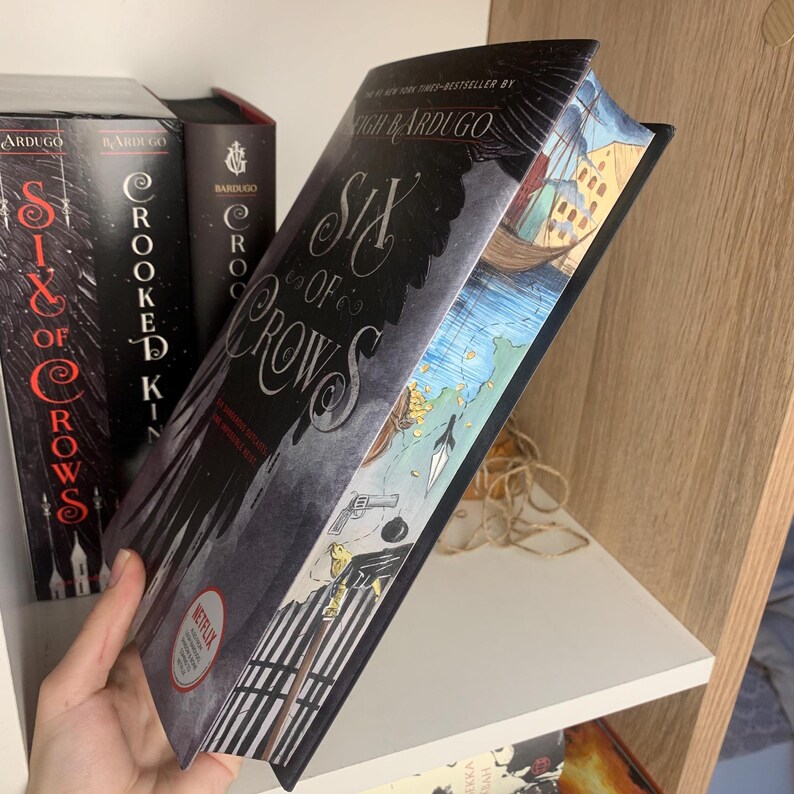 Może przedstawiać: Twarda oprawa książki zatytułowanej "Six of Crows" autorstwa Leigh Bardugo, z ciemną okładką z tytułem w eleganckiej czcionce. Krawędzie książki przedstawiają kolorową ilustrację. Inne książki są widoczne na p&oacute;łce.