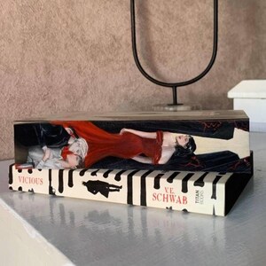 May include: A decorative wooden box featuring artwork from the book "Vicious" by V.E. Schwab. The box has a painted image of a woman in a red dress and a man in a suit. The book title and author's name are visible.