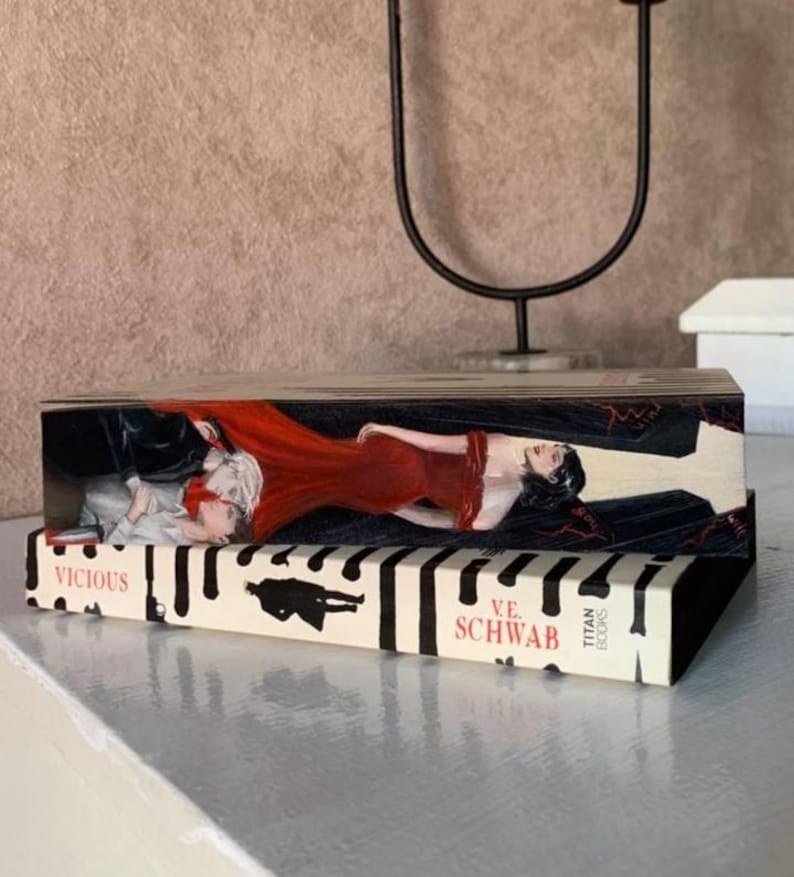 May include: A decorative wooden box featuring artwork from the book "Vicious" by V.E. Schwab. The box has a painted image of a woman in a red dress and a man in a suit. The book title and author's name are visible.
