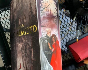 May include: A rectangular book titled ALCHEMISED by LinYu featuring a dark, gothic-style cover illustration of a castle and a side panel depicting a person with white hair in a grey cloak standing beside a person in a vibrant red dress.