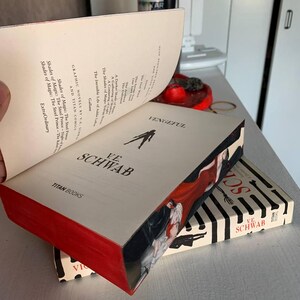 May include: A stack of books by V.E. Schwab, including "Vengeful" and "Shades of Magic." The books have red edges and feature illustrations of figures in red and black. The book covers are cream-colored with black and white striped designs.