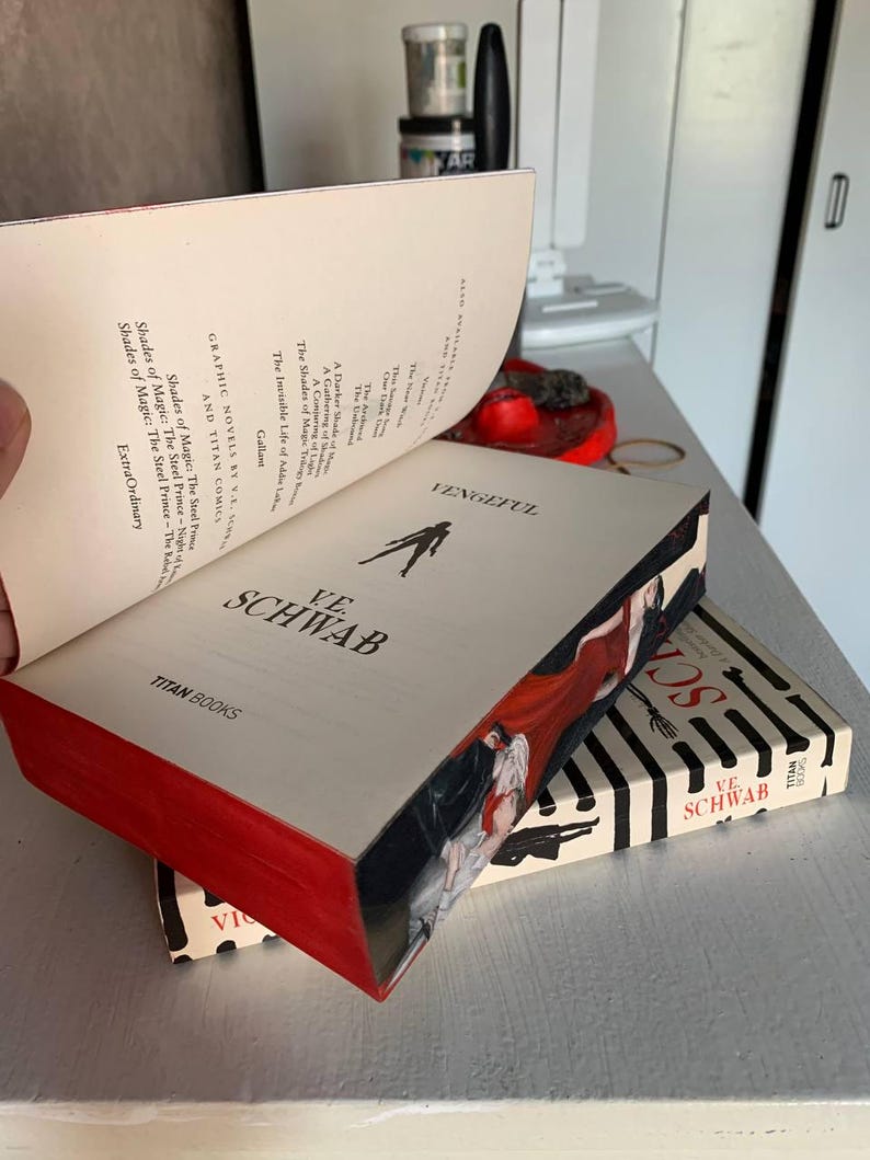 May include: A stack of books by V.E. Schwab, including "Vengeful" and "Shades of Magic." The books have red edges and feature illustrations of figures in red and black. The book covers are cream-colored with black and white striped designs.