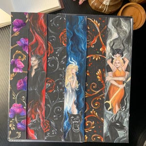 May include: A set of fantasy-themed bookmarks. Each bookmark displays a unique illustration on a dark background, with floral or ornate patterns, and characters with horns. The colour scheme includes purple, red, blue, and orange.