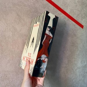 May include: A hardcover book titled "Vengeful" by V.E. Schwab. The cover features a woman in a red dress, and two men embracing. The spine has vertical black and white stripes. The book is held against a neutral background.