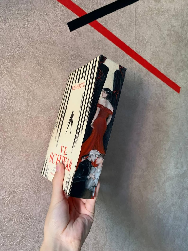 May include: A hardcover book titled "Vengeful" by V.E. Schwab. The cover features a woman in a red dress, and two men embracing. The spine has vertical black and white stripes. The book is held against a neutral background.