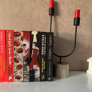 May include: A decorative black metal candle holder with two red candles, next to a row of books with colorful covers. The books include titles like "A Court of Thorns and Roses" and "Kingdom of the Wicked". The candle holder has a stone base.