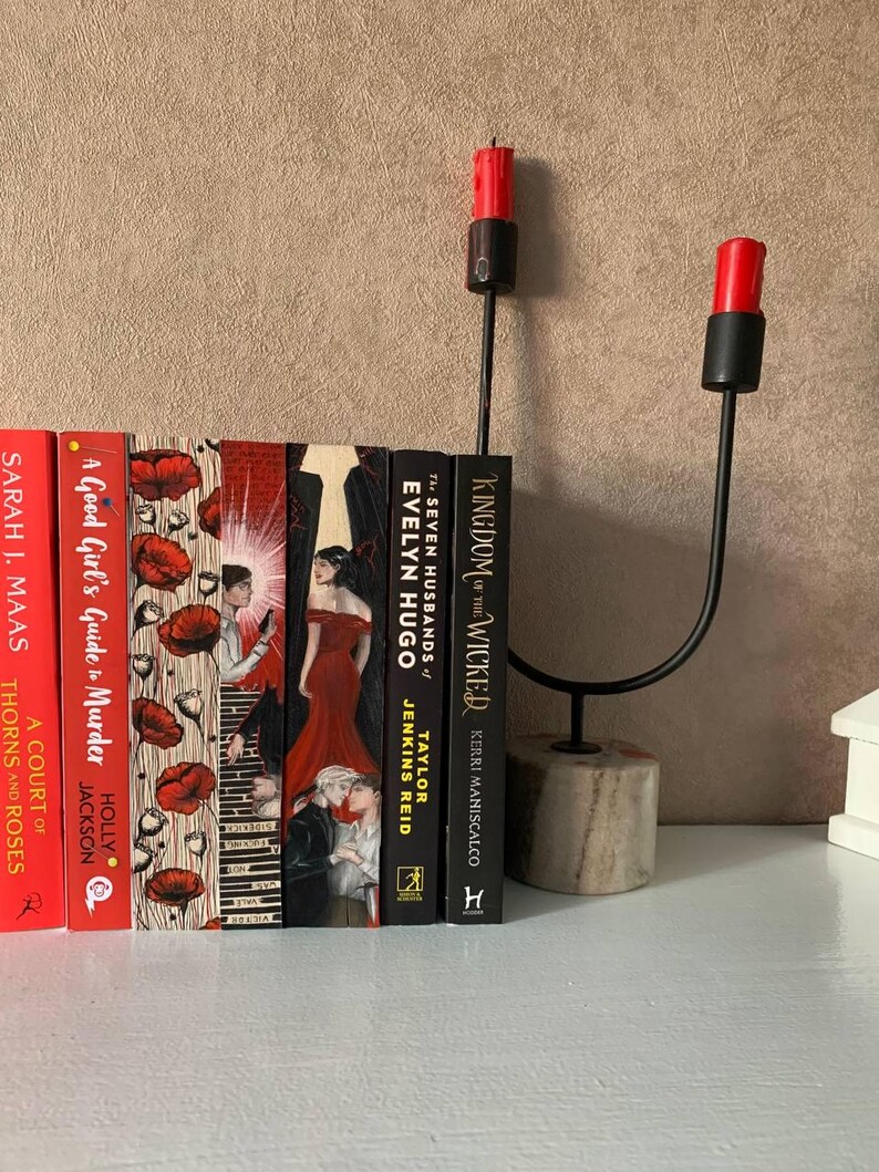 May include: A decorative black metal candle holder with two red candles, next to a row of books with colorful covers. The books include titles like "A Court of Thorns and Roses" and "Kingdom of the Wicked". The candle holder has a stone base.