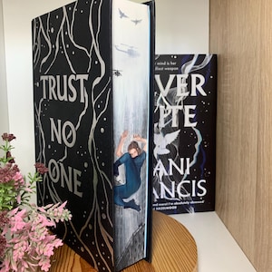 May include: A hardcover book with the title "TRUST NO ONE" in silver lettering on a black cover. The book's edge features an illustration of a person falling, with helicopters in the background. Another book with the title "VERITE" is visible.
