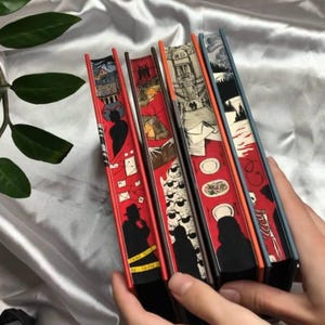 Puede incluir: Una colección de cinco libros de tapa dura con lomos ilustrados. Los lomos presentan fondos rojos con diversas ilustraciones en blanco y negro, incluyendo siluetas, dibujos arquitectónicos y comida.