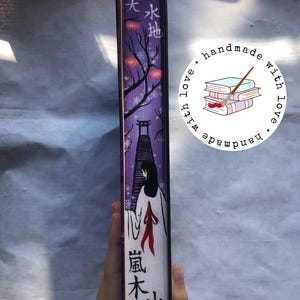 以下が含まれることがあります： 紫色の夜の風景が手描きされた本の背表紙。白い着物を着た人物、鳥居、赤い提灯が描かれています。背表紙には黒い日本語の文字と白い雪の結晶がデザインされています。handmade with loveと書かれた円形のステッカーが貼られています。