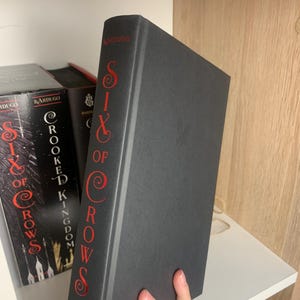 Może przedstawiać: Twarda czarna książka zatytułowana "Six of Crows" autorstwa Leigh Bardugo. Grzbiet książki ma czerwone litery. Nazwisko autora, Bardugo, jest wydrukowane na g&oacute;rze grzbietu.