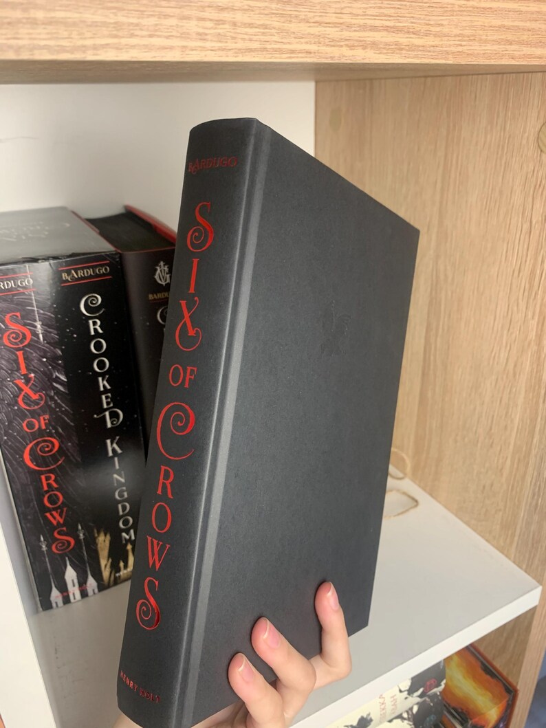 Może przedstawiać: Twarda czarna książka zatytułowana "Six of Crows" autorstwa Leigh Bardugo. Grzbiet książki ma czerwone litery. Nazwisko autora, Bardugo, jest wydrukowane na g&oacute;rze grzbietu.