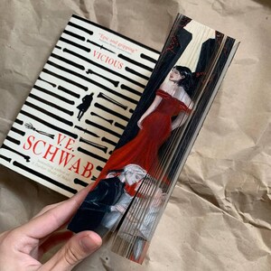 May include: A hardcover book titled "VICIOUS" by V.E. Schwab, with a black and white striped cover design. The book's pages are fanned out, revealing a painted illustration of a woman in a red dress and two men.