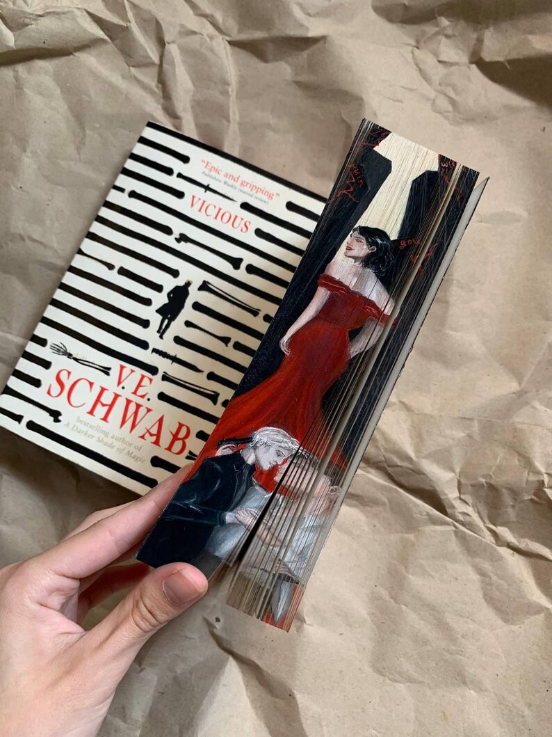 May include: A hardcover book titled "VICIOUS" by V.E. Schwab, with a black and white striped cover design. The book's pages are fanned out, revealing a painted illustration of a woman in a red dress and two men.