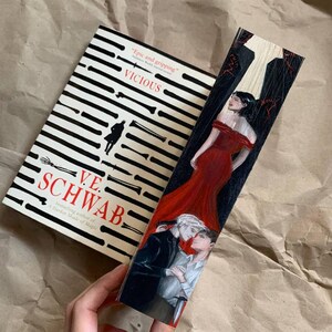 May include: A book titled "VICIOUS" by V.E. Schwab with a black and white striped design. A wooden bookmark with a painted illustration of two people embracing and a woman in a red dress.