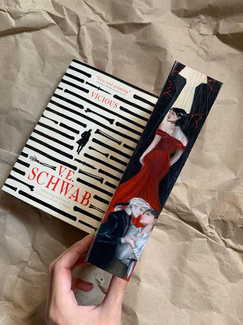 May include: A book titled "VICIOUS" by V.E. Schwab with a black and white striped design. A wooden bookmark with a painted illustration of two people embracing and a woman in a red dress.