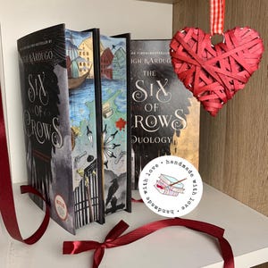 Może przedstawiać: Cztery książki, w tym "Six of Crows" i "The Six of Crows Duology", są wystawione na p&oacute;łce. Okładki książek przedstawiają ilustracje krajobrazu miejskiego, map i kruka. Czerwona ozdoba w kształcie serca wisi w pobliżu, a wstążka jest zawiązana w kokardę.