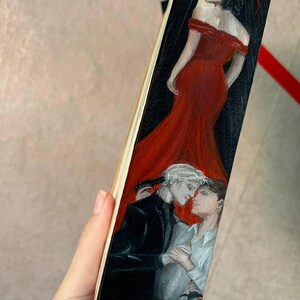 May include: A hand-painted wooden bookmark featuring a woman in a red dress and two men in a romantic embrace. One man holds a knife. The bookmark has a black and red background.