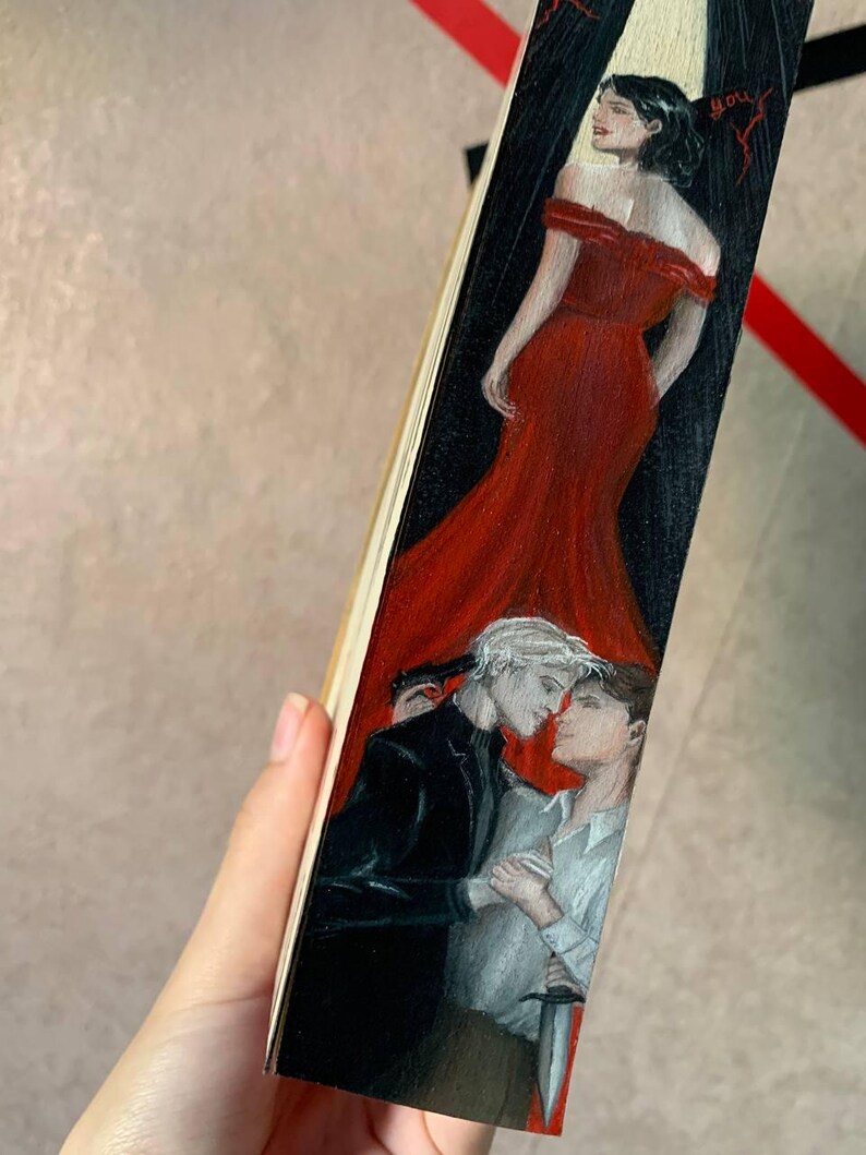 May include: A hand-painted wooden bookmark featuring a woman in a red dress and two men in a romantic embrace. One man holds a knife. The bookmark has a black and red background.