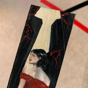 May include: A hand-painted wooden bookmark featuring a woman in a red dress. The bookmark has a black background with a white spotlight and red text that reads "will ruin you."