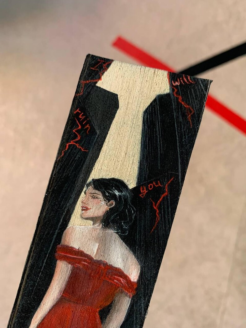 May include: A hand-painted wooden bookmark featuring a woman in a red dress. The bookmark has a black background with a white spotlight and red text that reads "will ruin you."